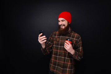 Photo of bearded hipster using mobile phone while holding cup with coffee standing over dark wall