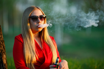 Vape woman. Young beautiful blonde girl in red dress and sunglasses smokes an electronic cigarette...