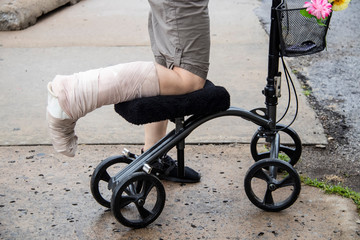 Woman with wrapped leg on steerable knee walker scooter with flowers on basket and mobile phone...