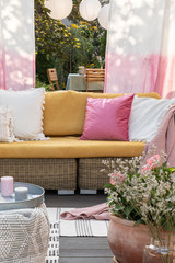 Flowers and candles in front of rattan sofa with colorful pillows on the terrace with drapes. Real photo