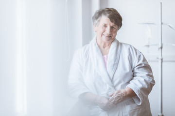 Senior grey lady wearing white bathrobe standing by the window at nursing home