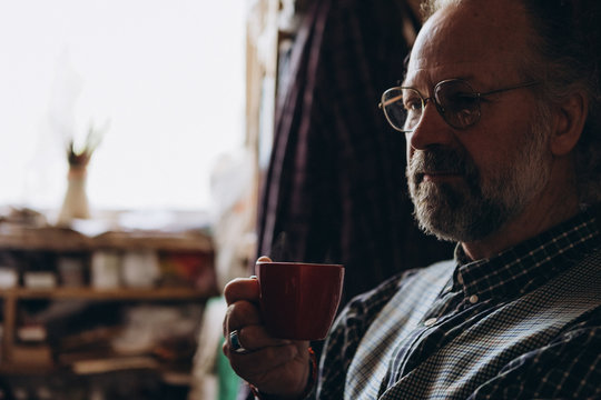 A stylish old painter relaxes after work in his own little cozy studio while sitting and drinking tea or coffee or alcohol near  window with day light