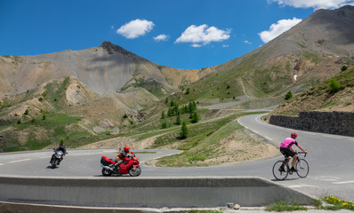Travelers on motorbikes and road bicycle ride up a switchback road in the Alps.