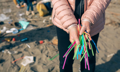 Unrecognizable woman showing handful of straws collected on the beach with group of volunteers...