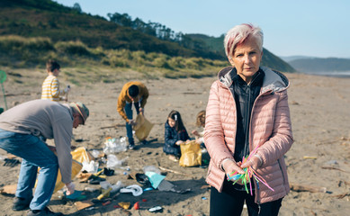 Serious woman showing handful of straws collected on the beach with group of volunteers working in...