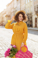 Outdoor portrait of young beautiful happy smiling lady wearing stylish yellow polka dot dress, straw hat, elegant wrist watch, holding straw wicker bag with peonies, walking in street of European city © Victoria Fox