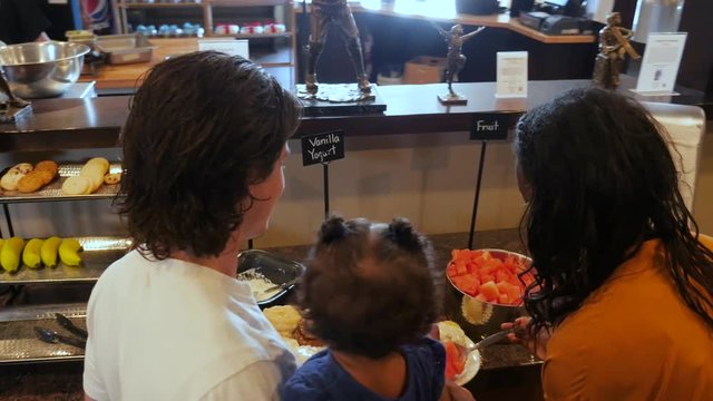 family with baby serve food from buffet line. A young multi-racial family dish up food down a breakfast buffet line. A young father holds his little girl in his arms while his wife fixes plates.
