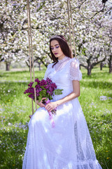  A young beautiful girl with a bouquet of lilac sits on a makeshift swing in an apple orchard in spring. White lace dress on the perfect figure of the model. Photo session in the open.