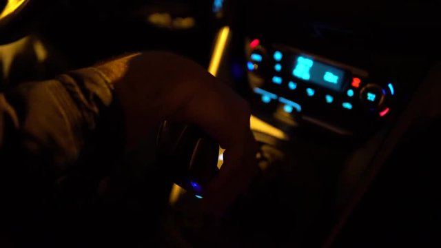 Driver man controls auto and hold hand on gear shifter or gear knob gear stick at night shadows road lights. Close up of hand and gear shift gear stick in car. Change gear in car. Night driving.