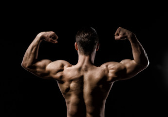 Muscular model sports young man on a dark background. Sporty healthy strong muscle guy showing his back and  big shoulders,biceps, triceps. Sexy male flexing his muscles. Workout bodybuilding concept.