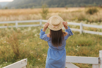 Back view of Pregnant woman in nature try on knitted hat. Background of field and white fance near the farm