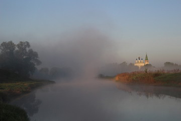 Misty cloud on river, green shore, trees, church in summer morning