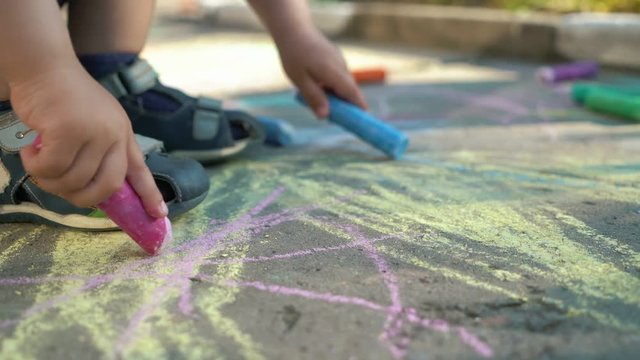 Little baby sitting in a shadow in sunny summer day and draws with crayons on asphalt road in the park. Colorful chalks in kid's hands. Close up