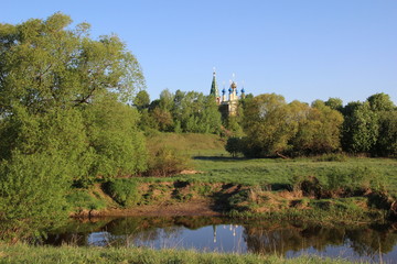 Sandy river beach in sunny summer day with green grass, trees, church under blue sky
