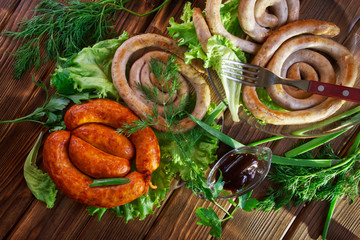 Grilled sausages and transparent sauceboat lie on a wooden surface in the sun along with greens: lettuce, dill, onion and parsley. Picnic - barbecue