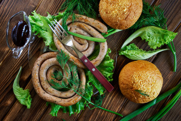 Grilled sausages, corn buns, transparent sauceboat and fork lie on a wooden surface with greens: lettuce, dill, onion and parsley. Picnic - barbecue