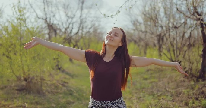 Happy brunette girl walks through the forest and enjoys the weather. Young woman raises her hands up and touches the branches of trees, she is pleased.