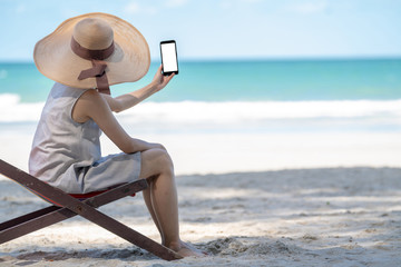 Fototapeta na wymiar Young adult woman traveler in mini dress sitting on beach chair on tropical island sand beach in summer day holidays vacation holding or using smartphone with blurred blue sea and beach backgrounds.