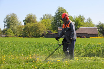 European senior man mowing the lawn with mower trimmer in a field on background of village houses on a Sunny summer day - working with garden tools