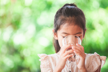 Cute asian child girl drinking fresh water from glass on green nature background