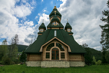 Fototapeta na wymiar Wooden green chapel on a hill in the forest. Rustic chapel. Church against the forest and sky. Orthodox, christian monastery in summer.