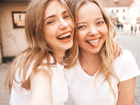 Two young smiling hipster blond women in summer white t-shirt clothes. Girls taking selfie self portrait photos on smartphone.Models posing on street background.Female shows tongue