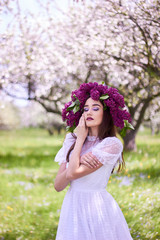 Portrait of a young beautiful girl in a wreath of lilacs. White lace dress on the perfect figure of the model. Photo session in the spring garden in the open.