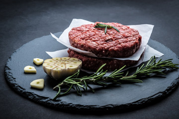 Raw burger beef patty with rosemary and garlic on dark cooking background