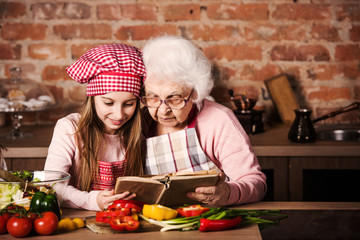 Little granddaughter reading recipe book with her granny sitting at kitchen