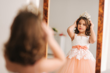 Naklejka premium Little beautiful girl with brown hair in a Peach-colored dress. Girl try on diadem. Happy little princess look in to the mirror