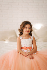 Naklejka premium Little beautiful girl with brown hair in a Peach-colored dress. Girl sitting on the sofa