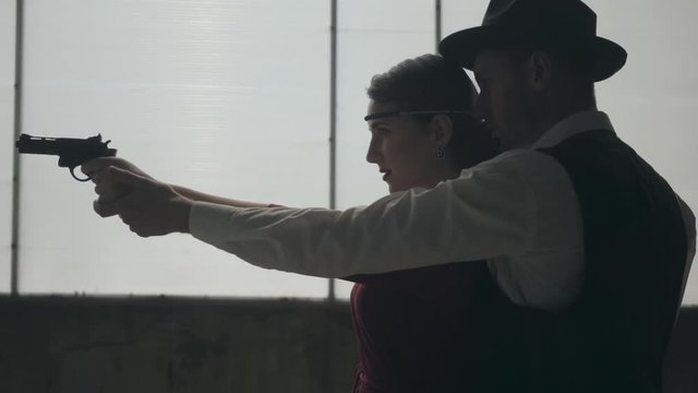 Portrait elegant young woman in red dress aiming a gun, handsome gallant man stands behind, directing her hand, side view. Happy couple of gangsters. Bonnie and Clyde