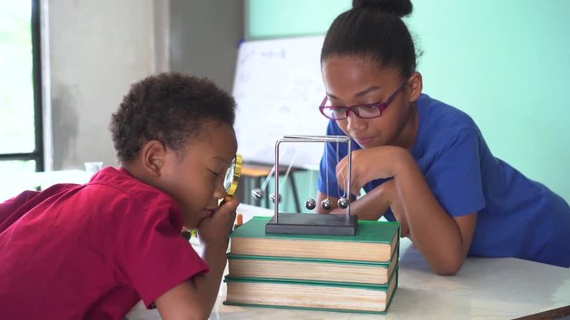 Two African American mixed kids using a magnifying glass and pulling a newton's cradles ball swing in science classroom - fun learning education concept