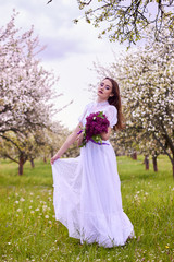 Obraz na płótnie Canvas Young beautiful girl with a bouquet of lilac in an apple orchard in spring. White lace dress on the perfect figure of the model. Photo session in the open.