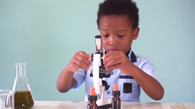 Young African American kid using microscope and experimenting scientific lab along with chemical substance tubes and flasks in classroom - science and learning education concept