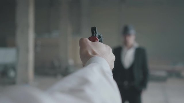 The hand of an unrecognizable man aiming a gun, blurred figure of a confident man in the suit and hat in front of him. The guy is going to kill his enemy. Mafia disassembly
