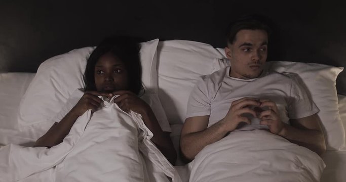 Young multi-ethnic couple lying in bed at night. Embarrassed and shy young couple before first intimacy in bed.