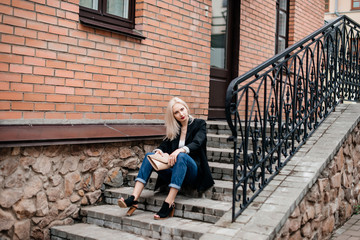 Fototapeta na wymiar Stylish girl in a black jacket and blue jeans sitting on the stairs outside