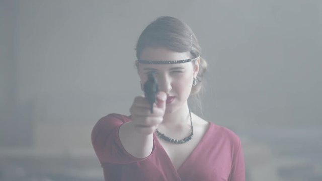 Confident attractive woman in stylish red dress is aiming a pistol at the camera in an abandoned dusty building, then lowers the gun. Dangerous lady. Retro, vintage style