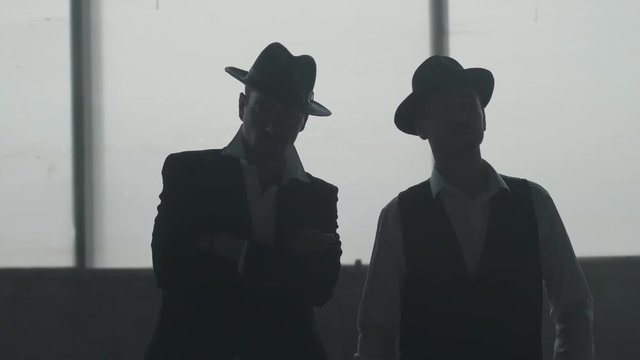 Two dark figures of confident well-dressed men in hats standing near each other in front of large window. Simultaneously both people putting hats on heads. Criminal authority, mafia