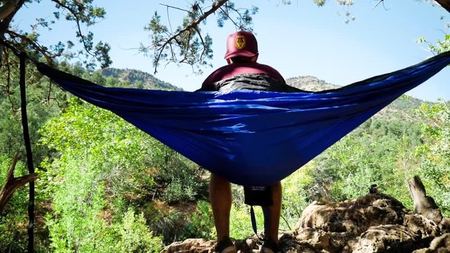 Guy wearing hat sitting in a blue hammock stretched between two short trees rocking back and forth looking into the distance with mountains and green trees in the background. Push in and tilt up.