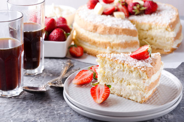 Closeup of a slice of sponge cake on the plate, glasses of coffee, bowl of strawberries, whole sponge cake on the background