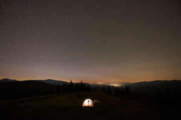 Summer camping at night. Tourist hikers tent brightly lit from inside near burning campfire on green grassy valley under dark blue starry sky.