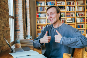 Joyful young man student working on a computer studying in the library on the background of the bookshelf. The guy shows a gesture class, rejoices success.