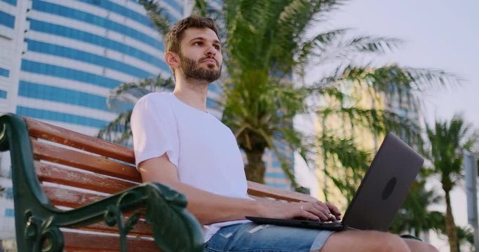 A young man in the summer of palm trees sitting with a laptop and typing on the keyboard