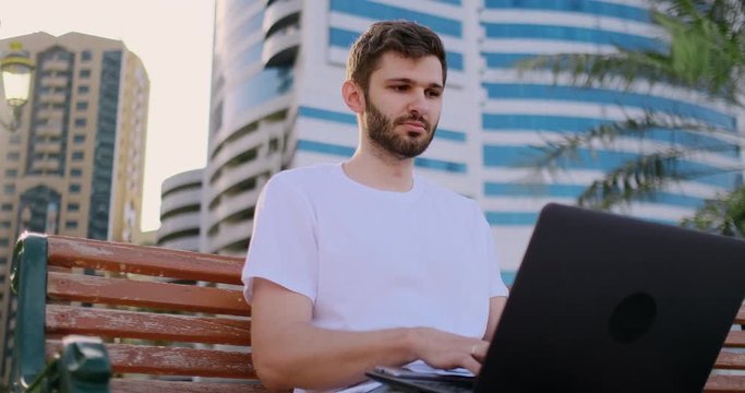 A young man in the summer of palm trees sitting with a laptop and typing on the keyboard