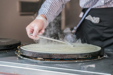 Making of crepes pancakes in open market festival fair. A hand is making crepes outdoors on a metal griddle with wooden stick on a outdoor summer fest