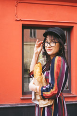 Young long-haired brunette woman in a bright dress against a red wall with bread baguettes in a paper bag