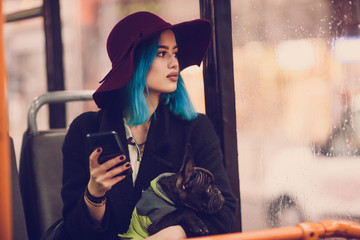 Young hipster woman sitting in city bus with her french bulldog and using mobile phone