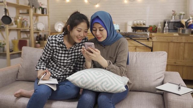 group of asian sisters shopping online christmas sale on smartphone. islam girl with blue headscarf share cell phone with chinese friend while writing on book doing homework. female point screen talk